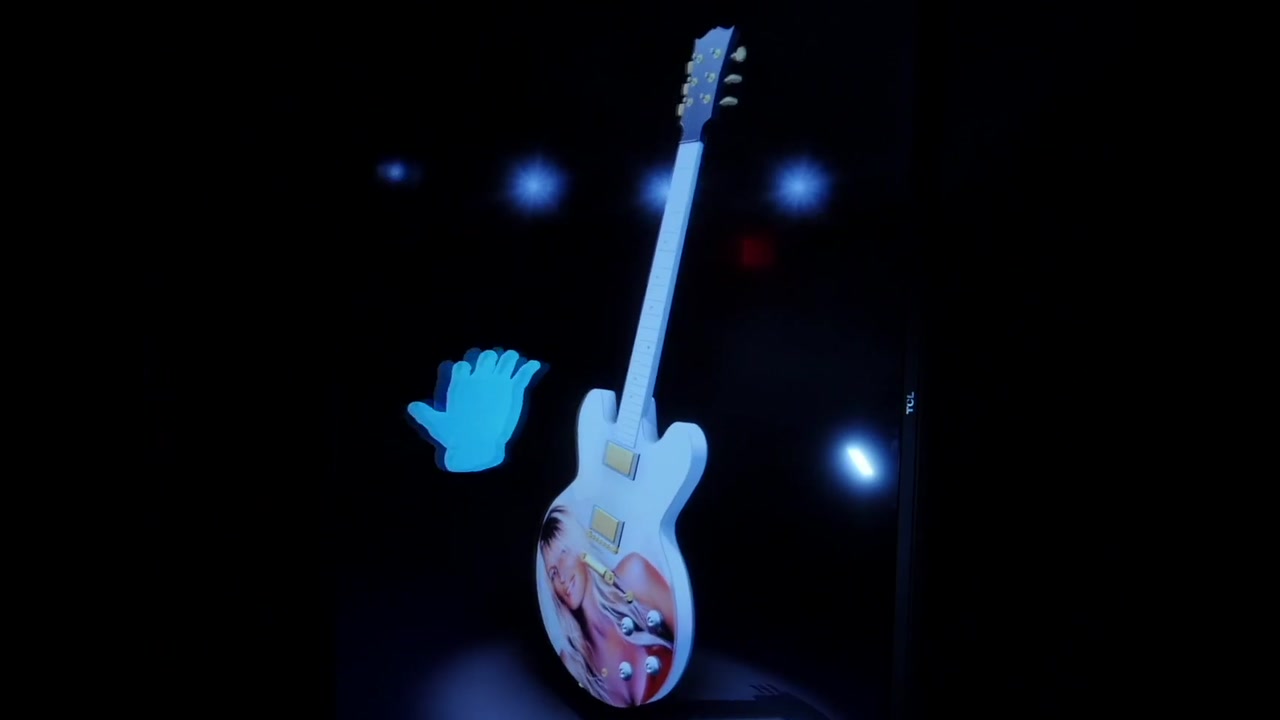 A hand shape floating beside a holographic guitar in a HoloTube, following a visitor’s hand in mid-air