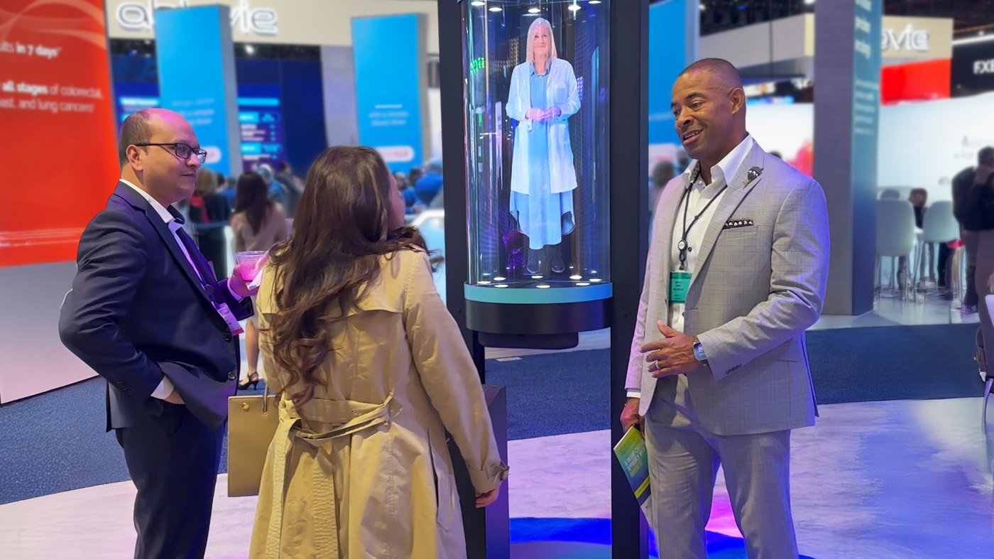 A sales representative talking with two visitors beside a HoloTube HalfPipe hologram host on a congress floor